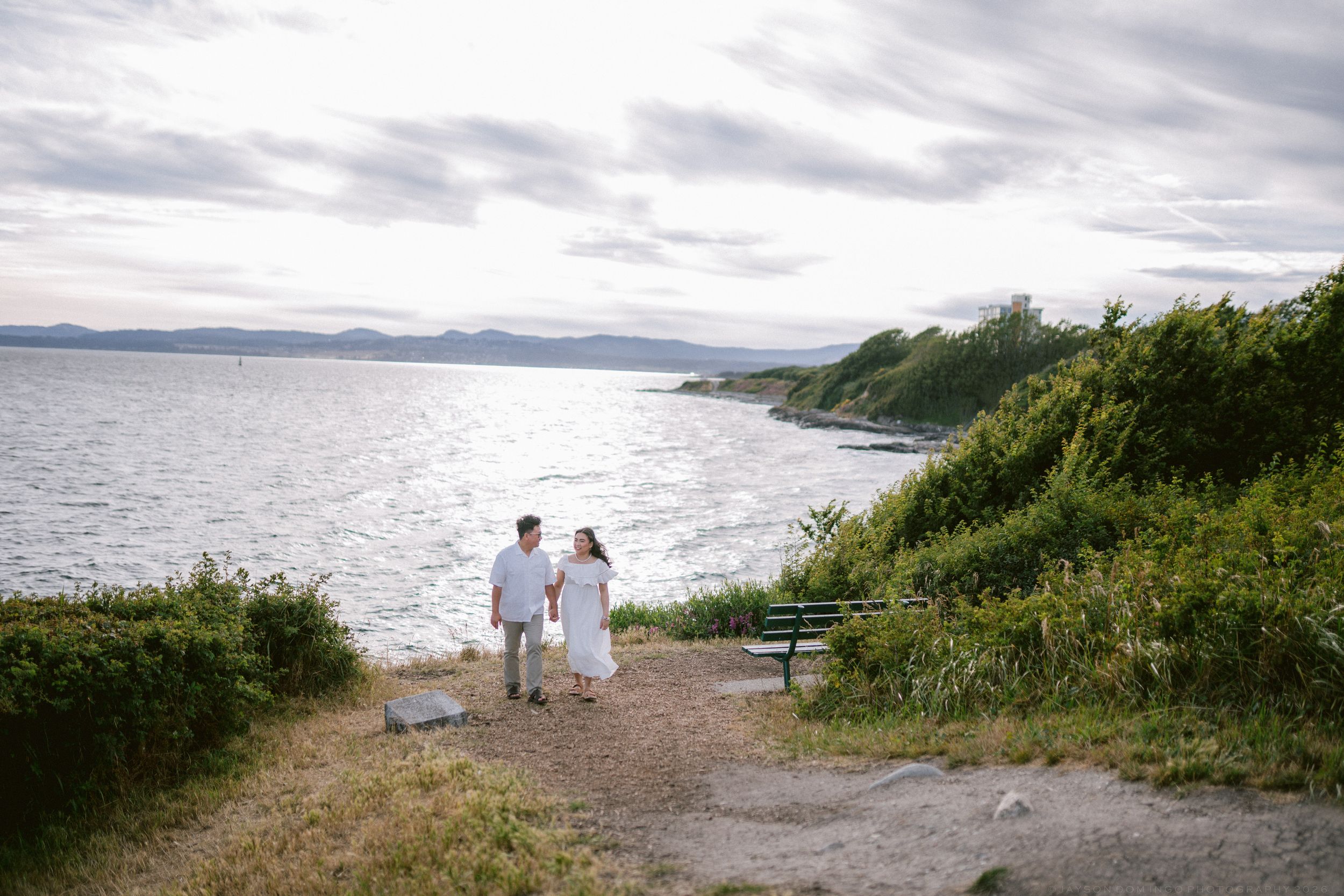 Couple in white outfits hold hands on a scenic coastal path with ocean views and lush greenery.