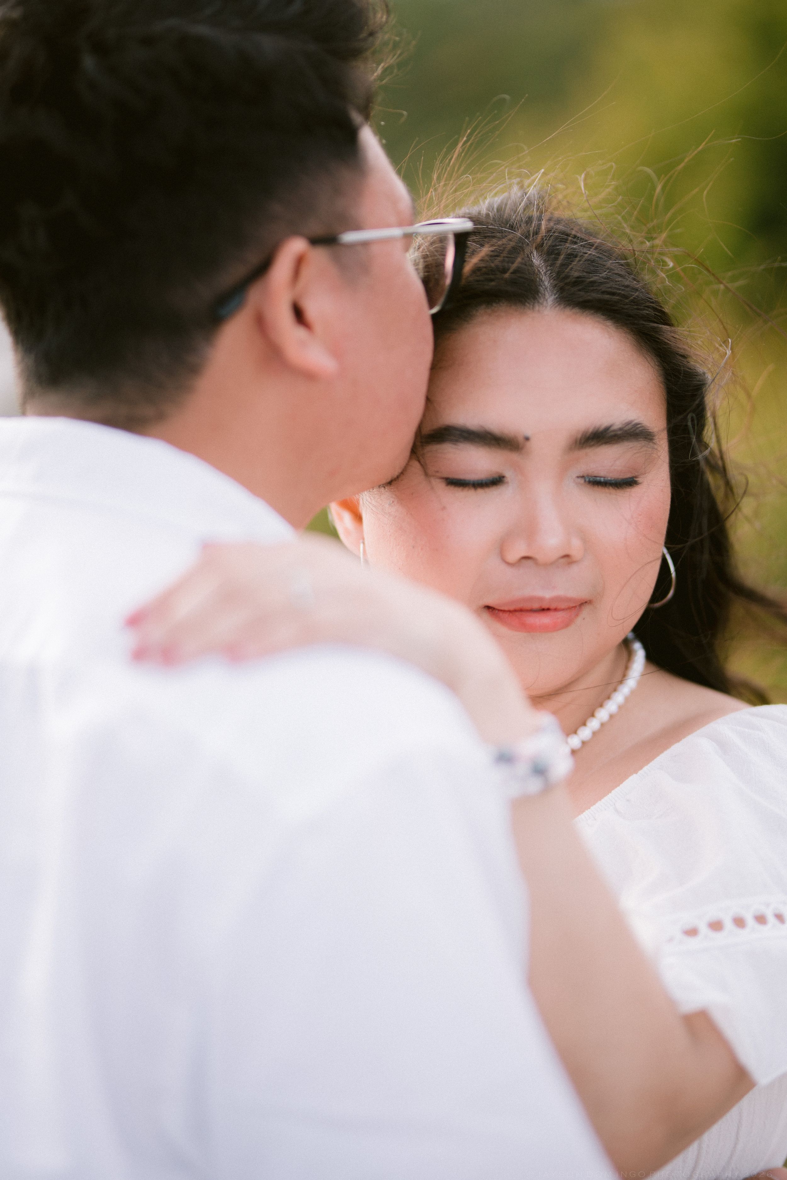 Close-up of a couple embracing tenderly, woman in white off-shoulder dress with pearl necklace, eyes closed peacefully.