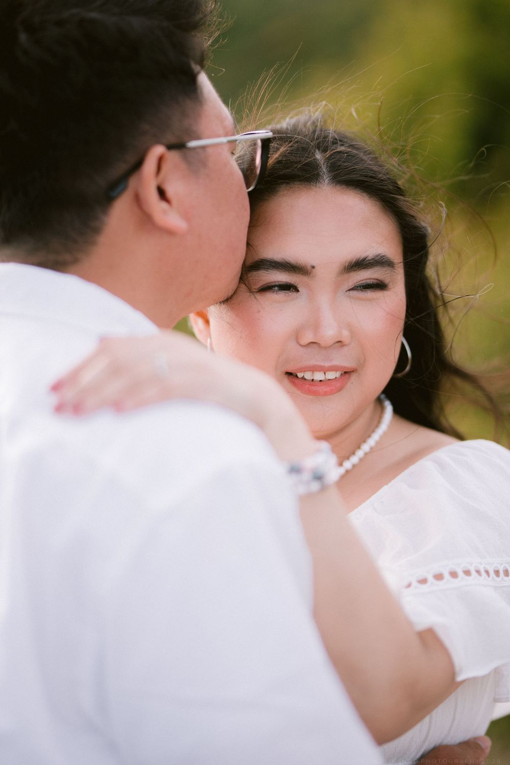 A smiling Asian woman in white dress with pearl necklace embraces a man in glasses outdoors.