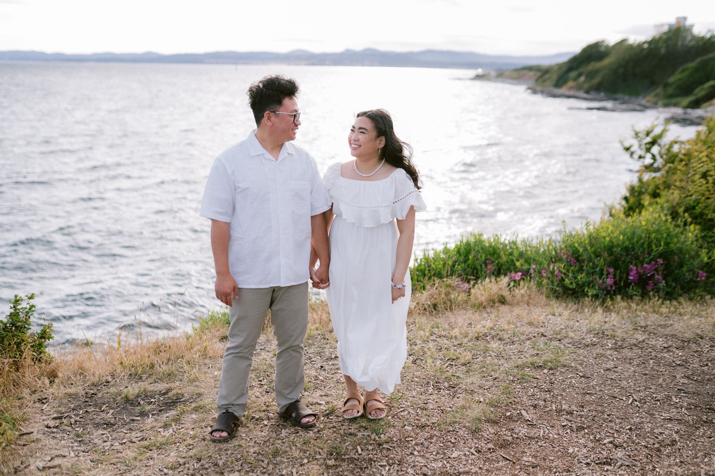 Happy couple holding hands by the seaside, woman in white dress and man in white shirt, smiling at each other.