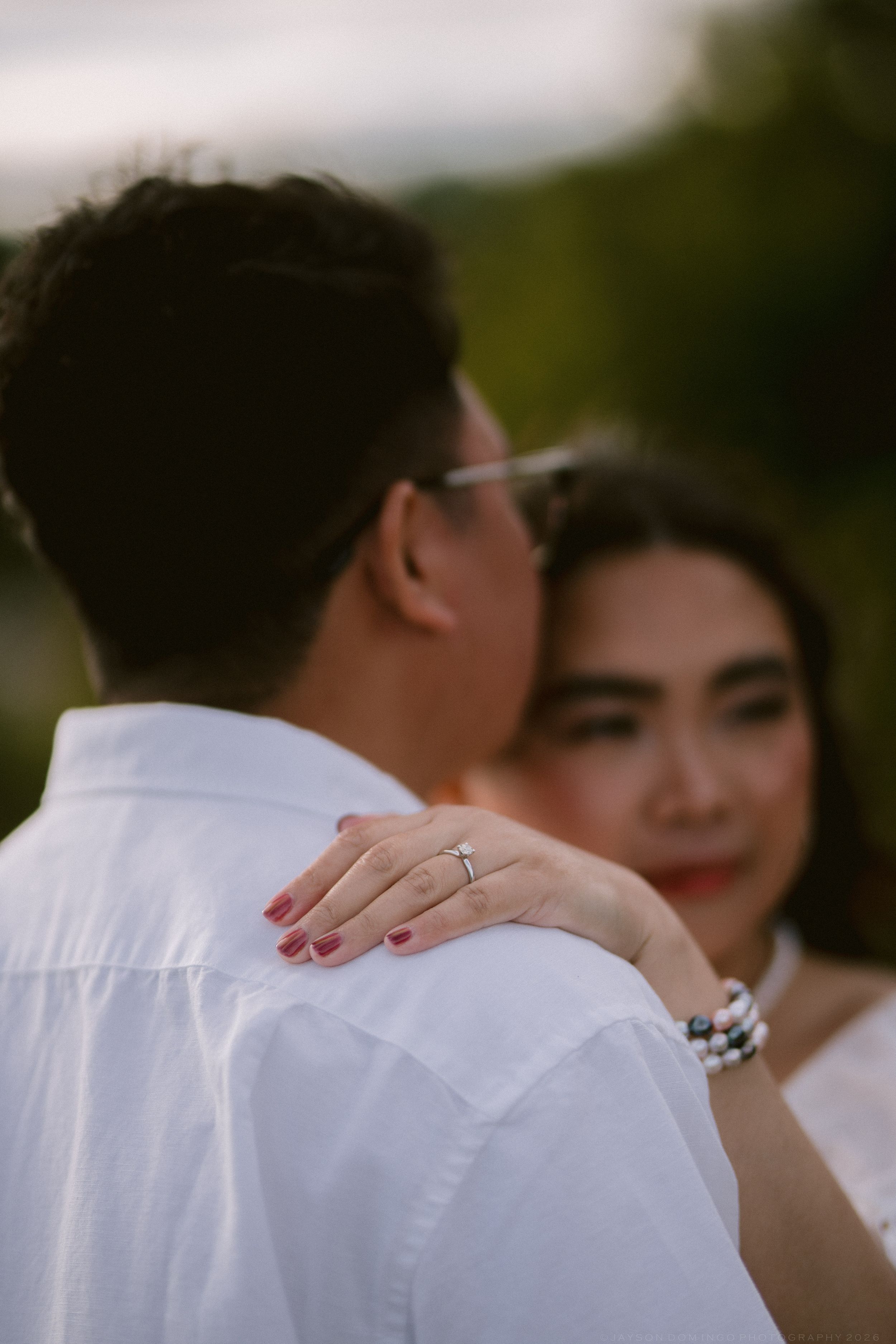 Close-up of woman's hand with engagement ring resting on man's white shirt shoulder during romantic outdoor couples session.