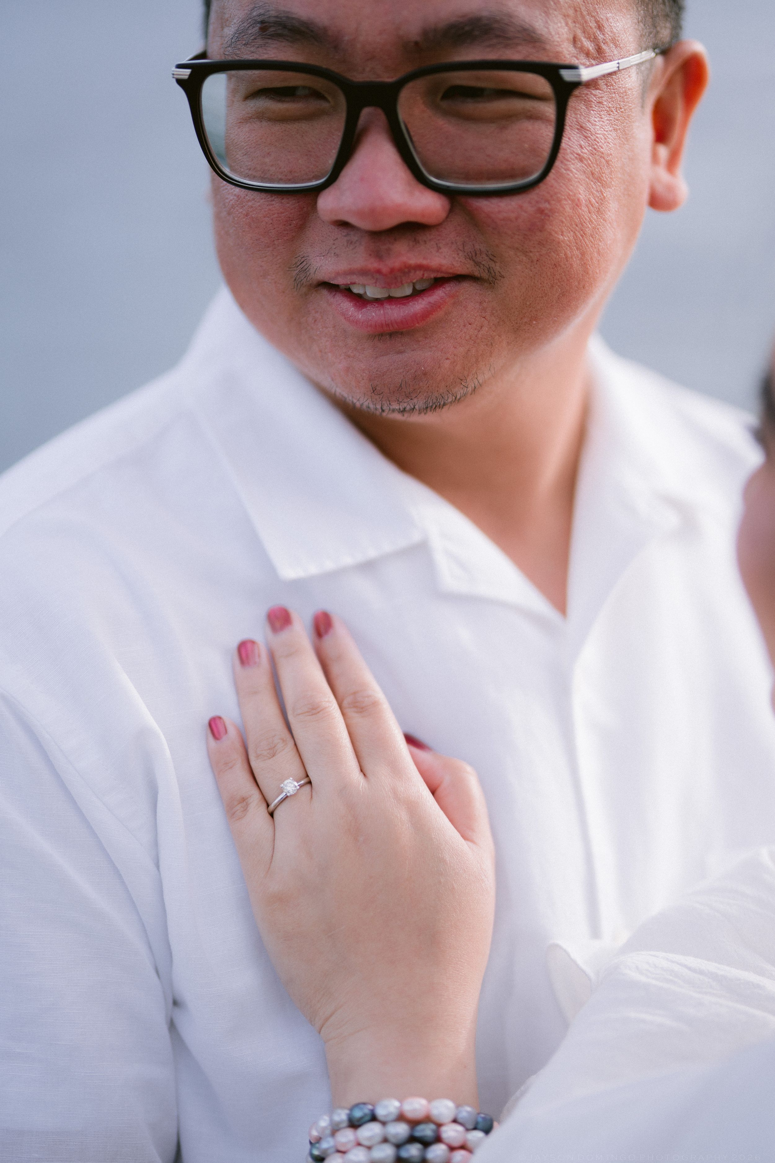 Man in white shirt with glasses smiles as a woman's hand with pink nails and ring rests on his chest.