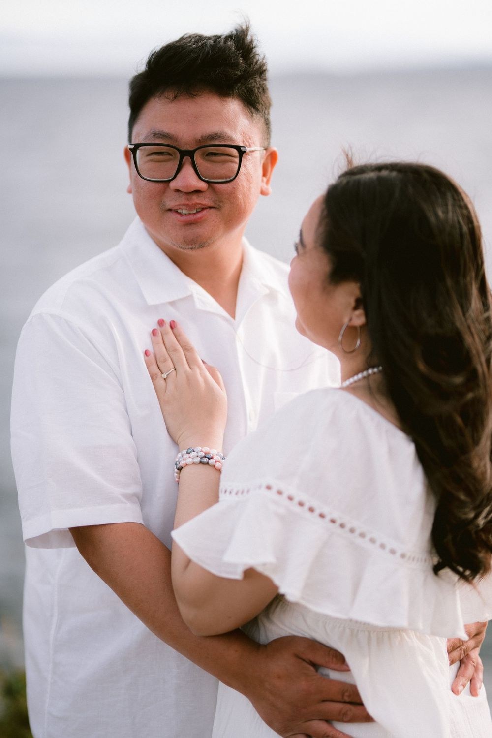 Couple in white outfits embracing outdoors, woman showing engagement ring, ocean in background.
