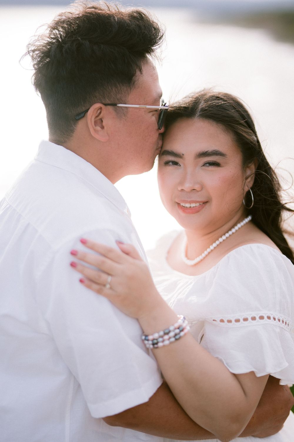 Asian couple embracing closely outdoors, woman smiling wearing pearl necklace.