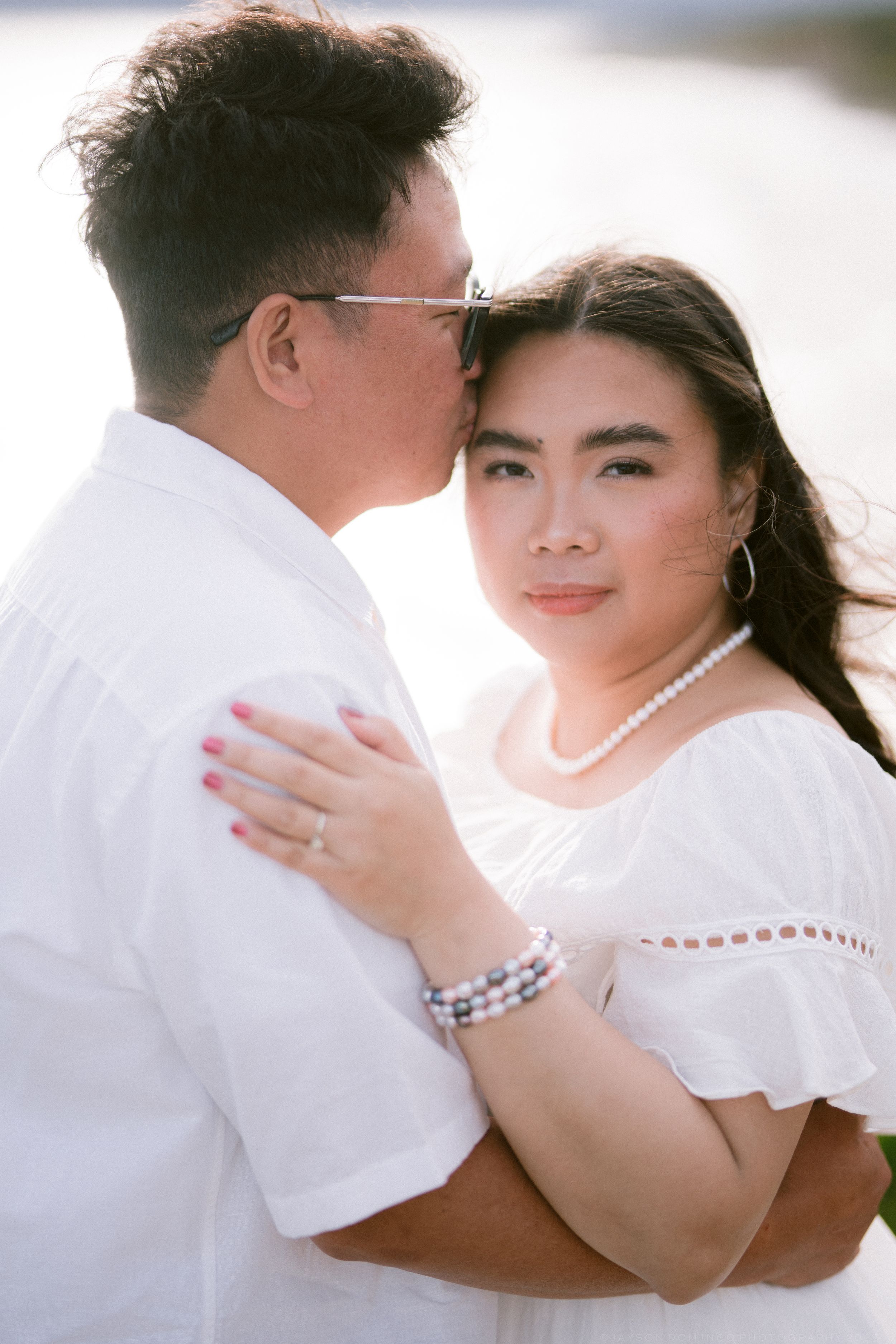 Asian couple in white sharing intimate moment outdoors in bright sunlight.