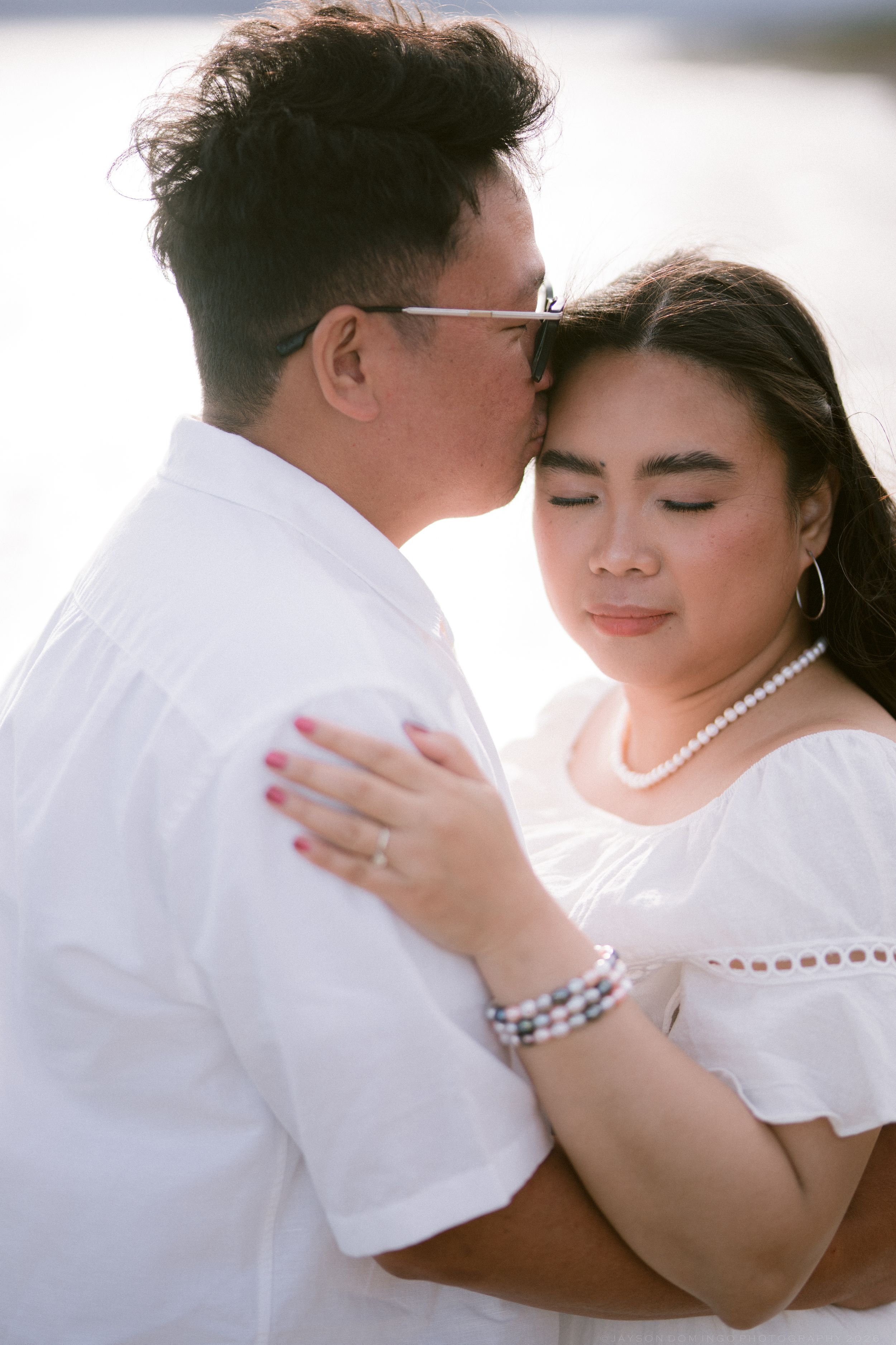 A tender moment as a man in glasses kisses a woman's cheek, both dressed in white near water.