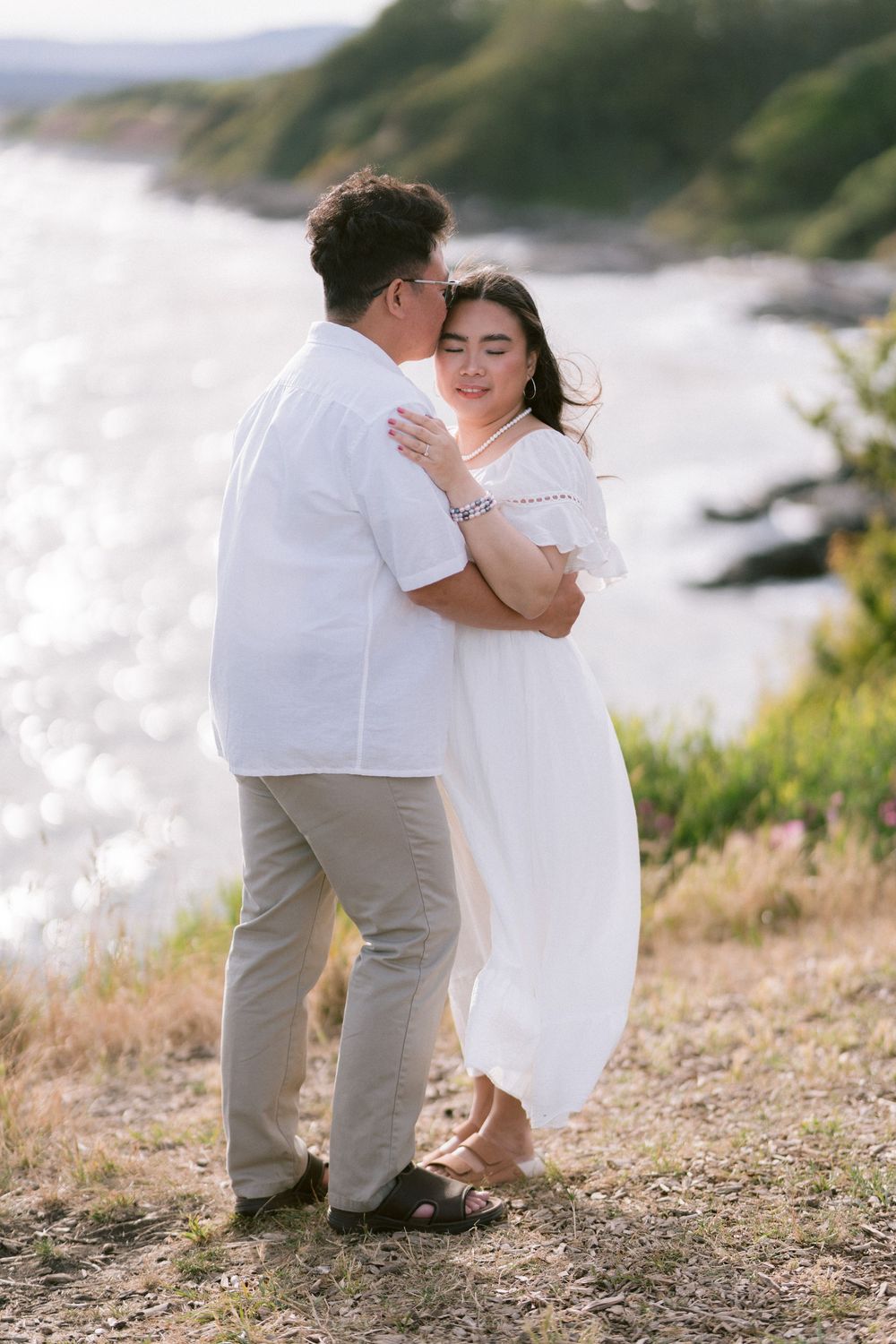 Couple embracing on a scenic coastal hillside, woman in white dress and man in white shirt, with sparkling water in background.