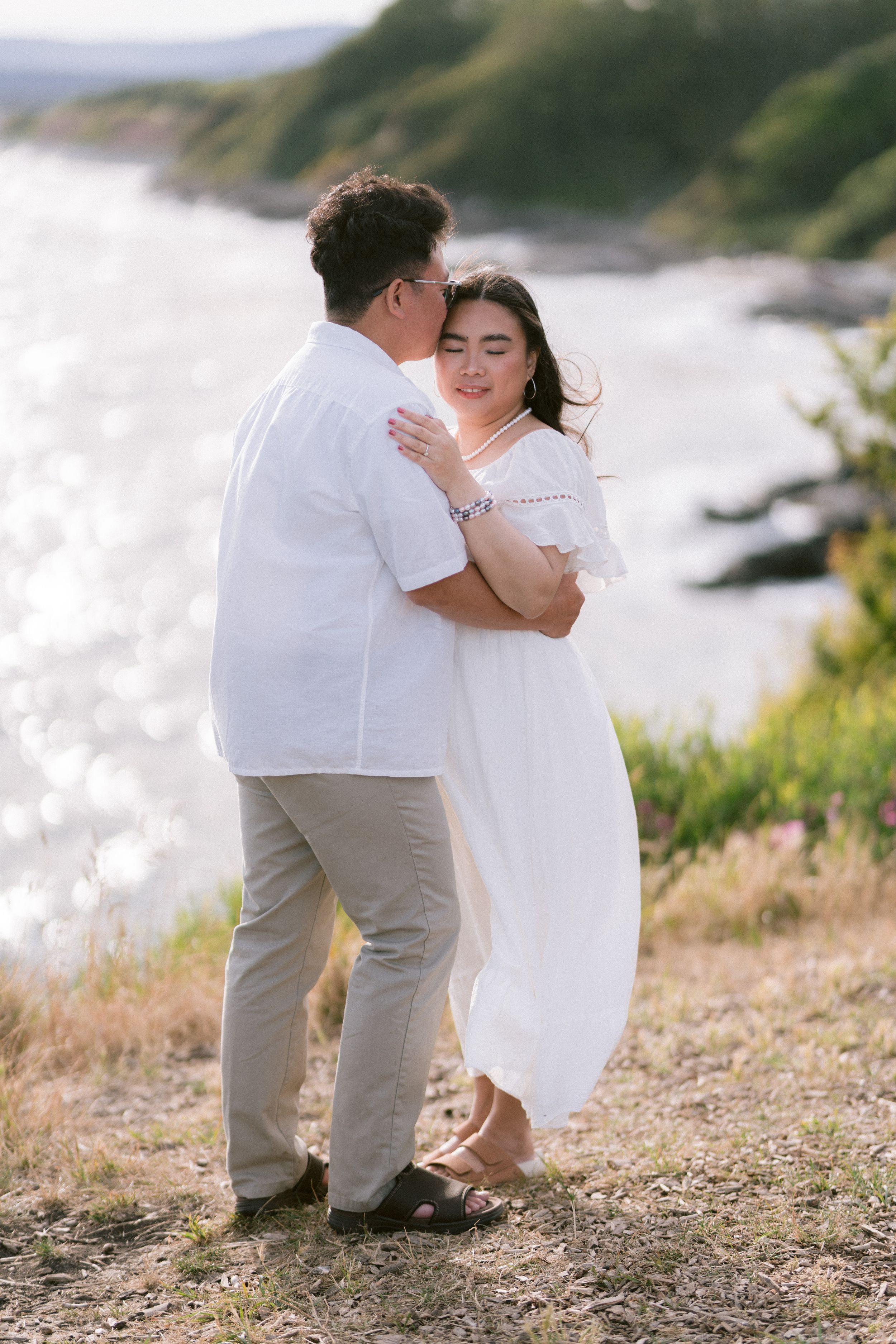 Couple embracing on a scenic coastal hillside, woman in white dress and man in white shirt, with sparkling water in background.