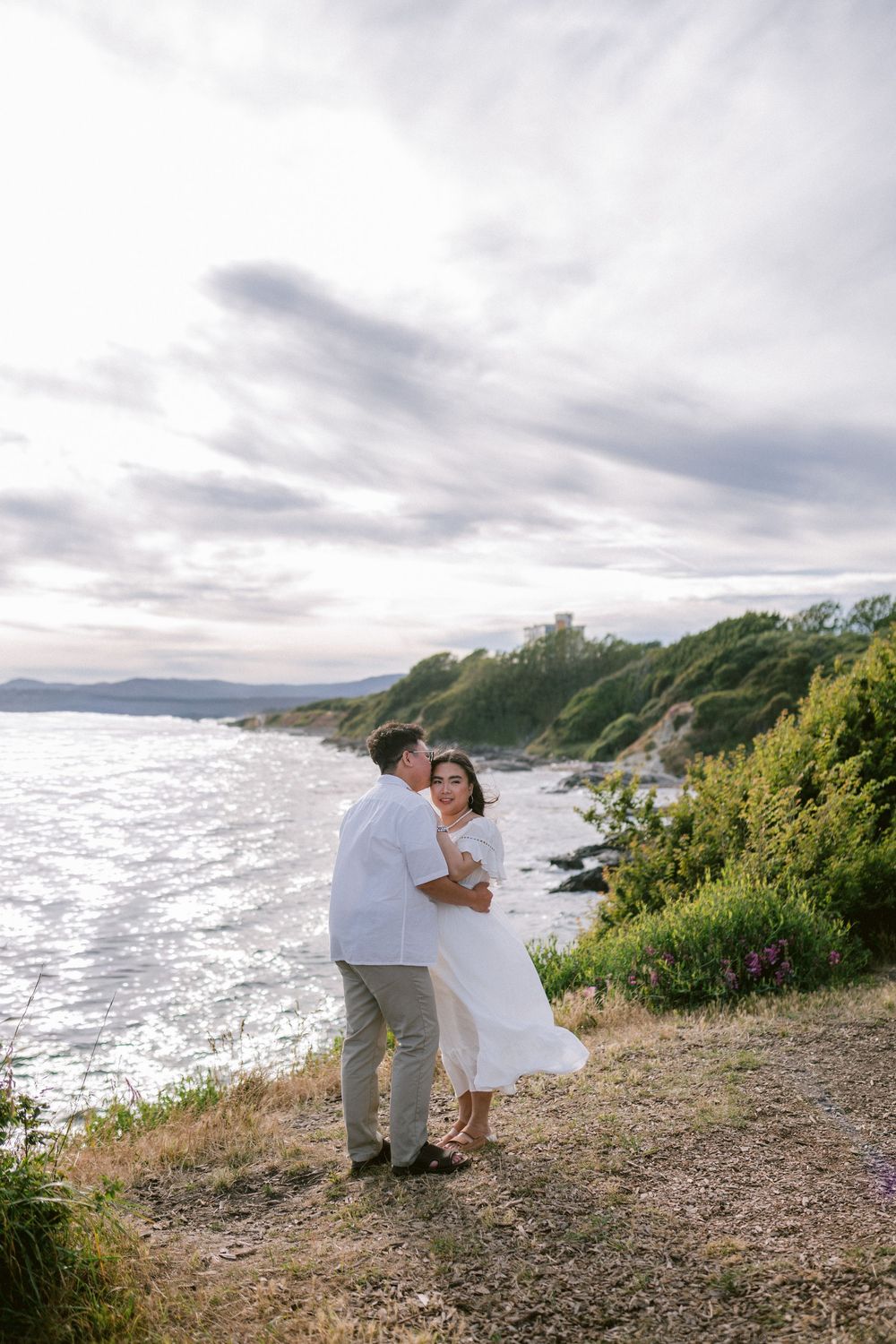Couple embracing on coastal cliff with ocean and green hills backdrop at dusk.