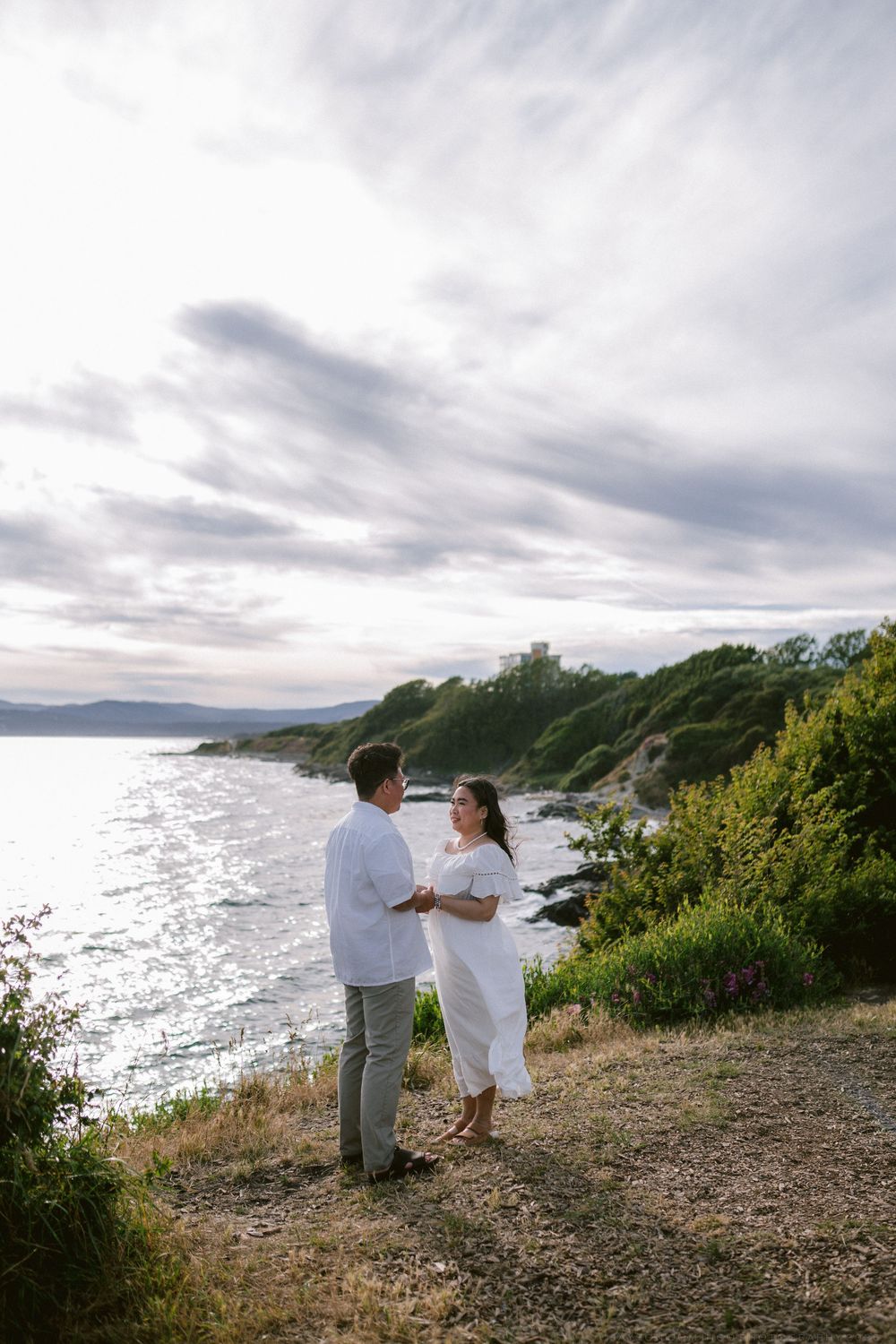 Couple in white outfits holding hands on scenic coastal cliff at sunset.
