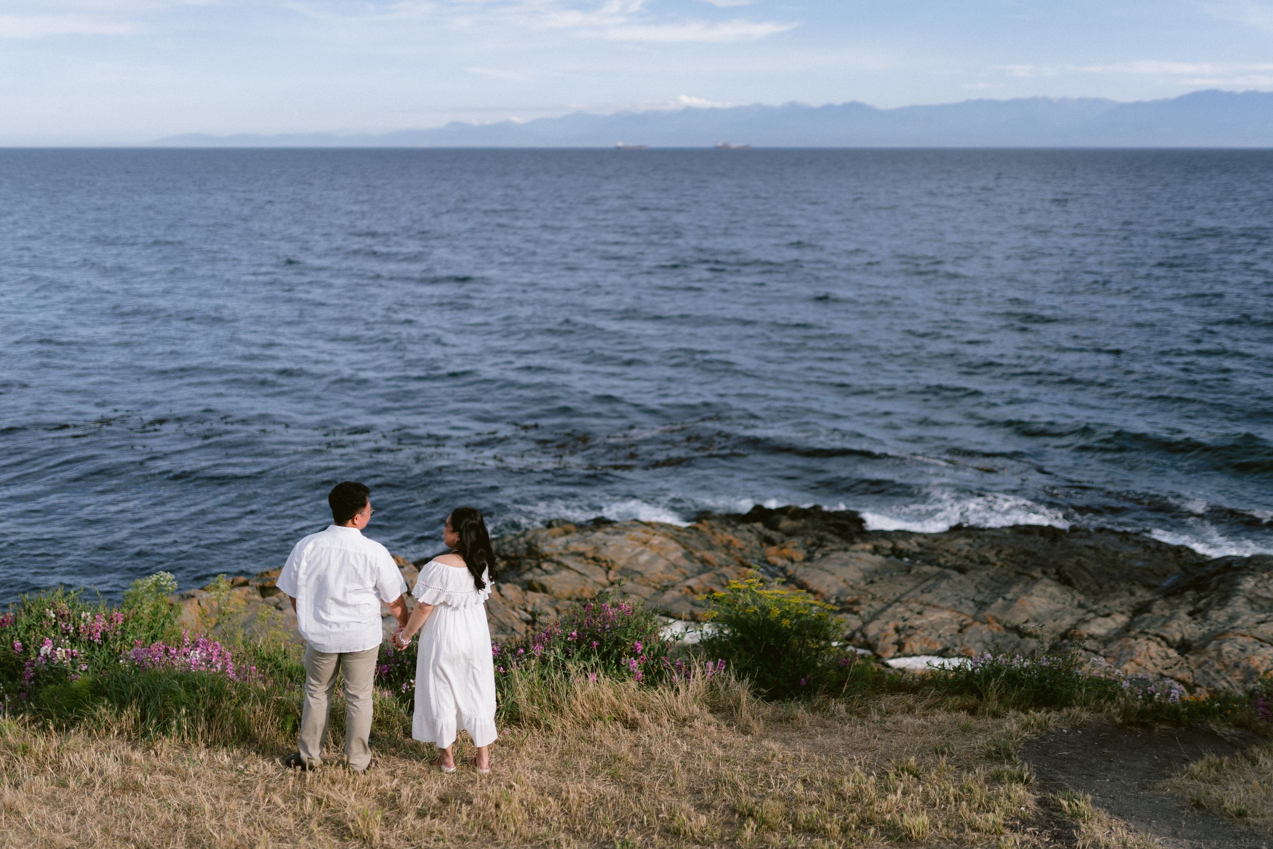 Couple in white standing on rocky coastal cliff overlooking calm blue ocean waters with distant mountains.