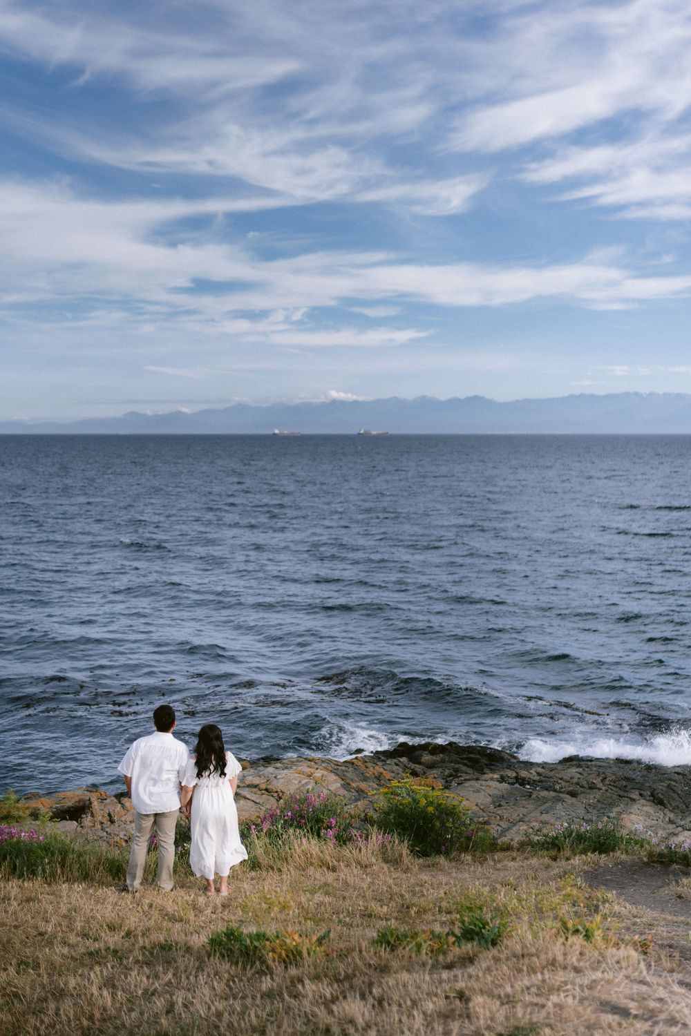Couple in white outfits holding hands while standing on rocky shoreline overlooking calm blue ocean with mountain backdrop.
