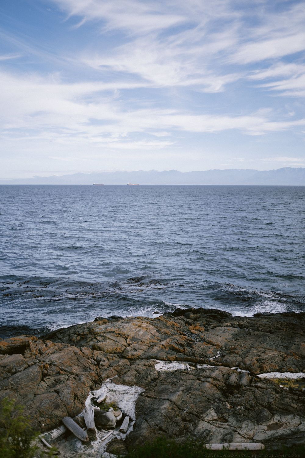 Rocky coastline with calm blue ocean waters and distant mountains under a partly cloudy sky.