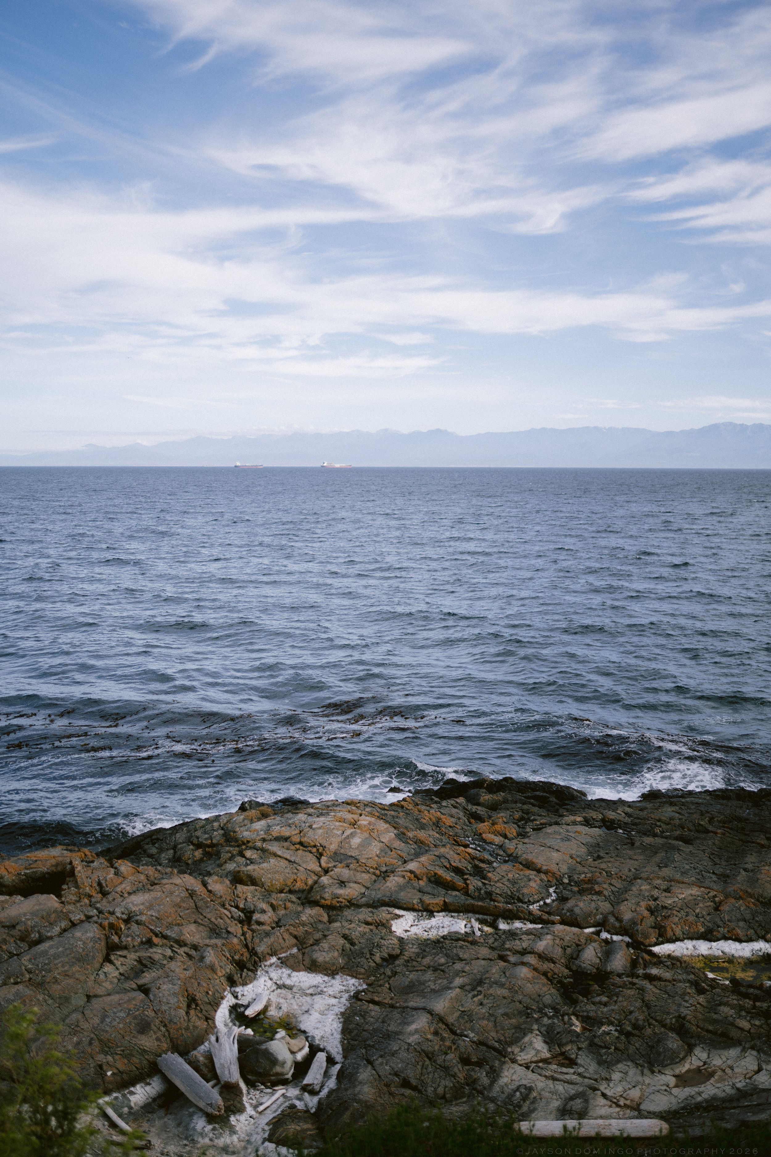 Rocky coastline with calm blue ocean waters and distant mountains under a partly cloudy sky.