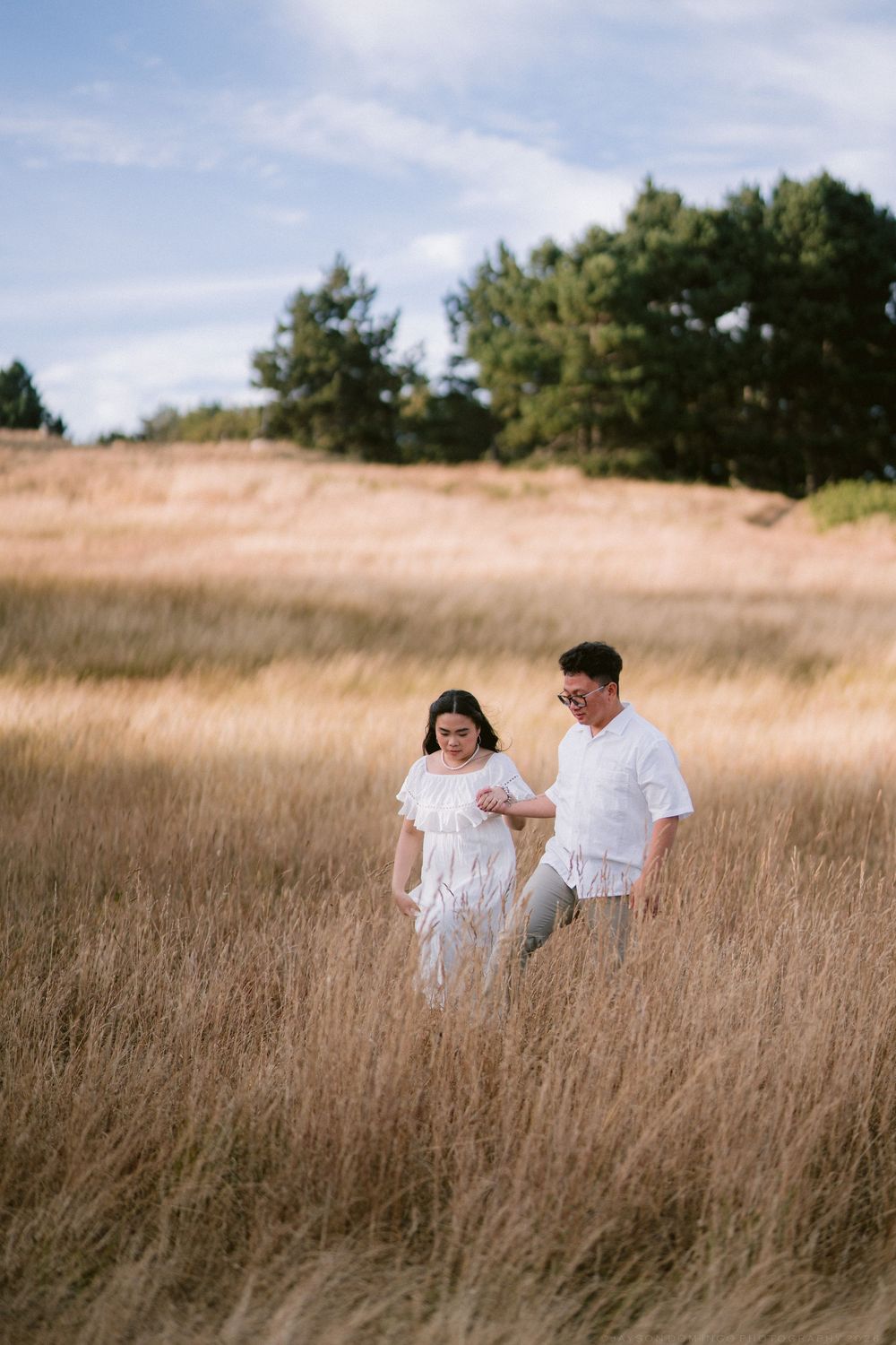 Couple in white outfits walking through golden wheat field with pine trees in background on cloudy day.