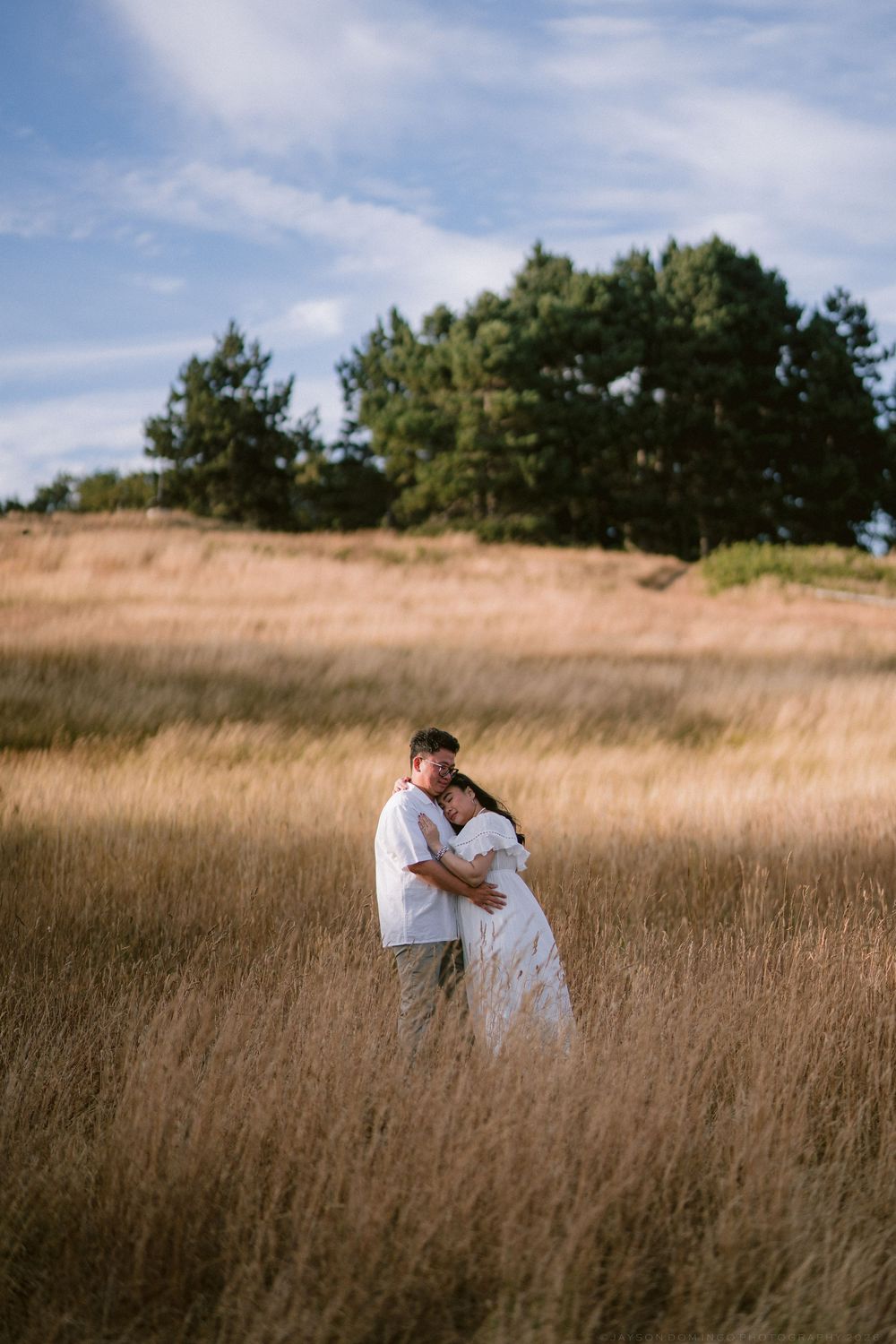 Romantic couple embracing in a golden wheat field with trees and blue sky, both dressed in white outfits.
