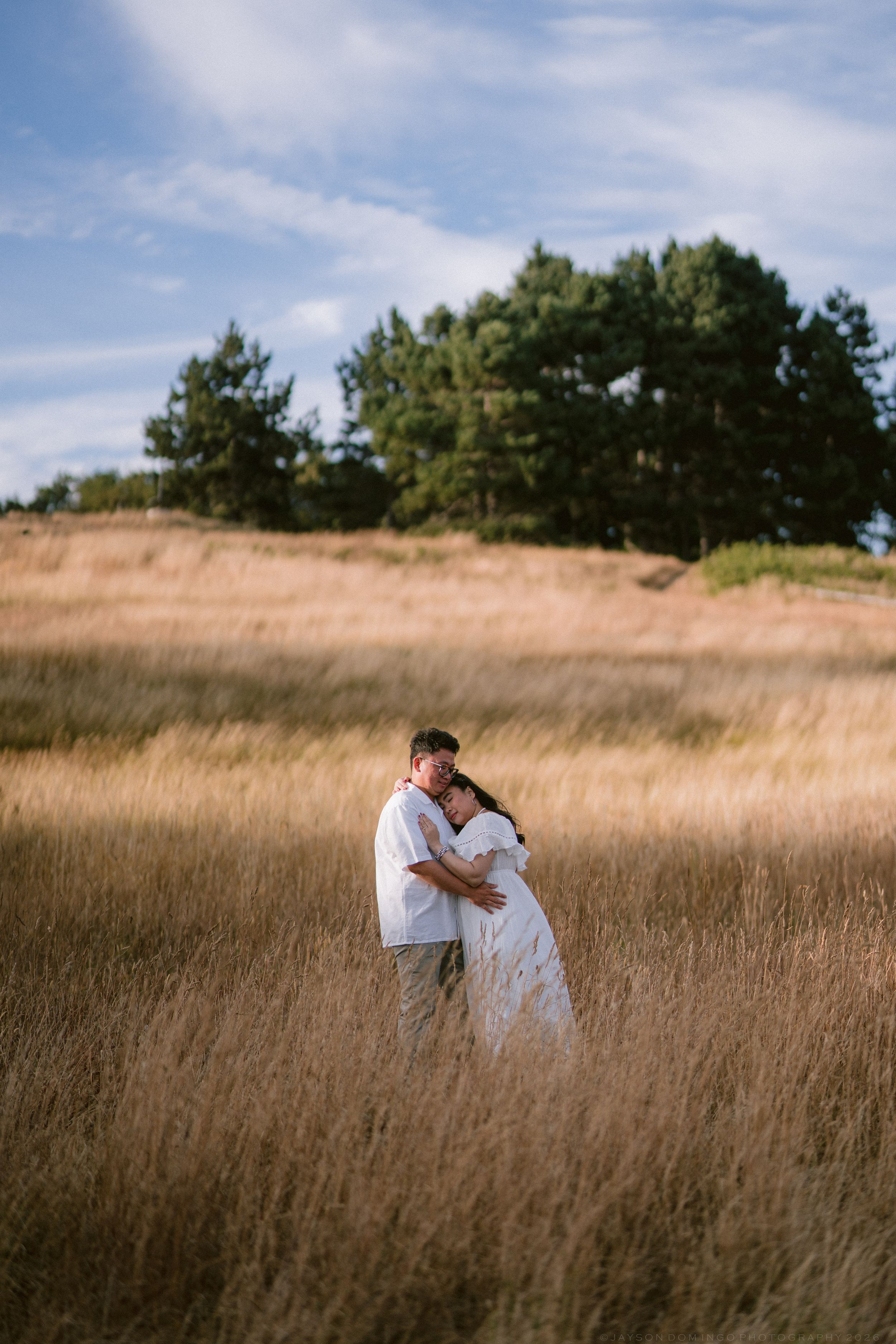 Romantic couple embracing in a golden wheat field with trees and blue sky, both dressed in white outfits.
