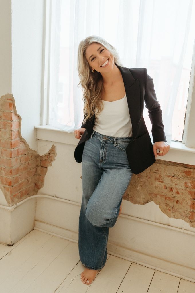 Smiling woman in black blazer, white top, and wide-leg jeans leaning against a window with exposed brick wall.