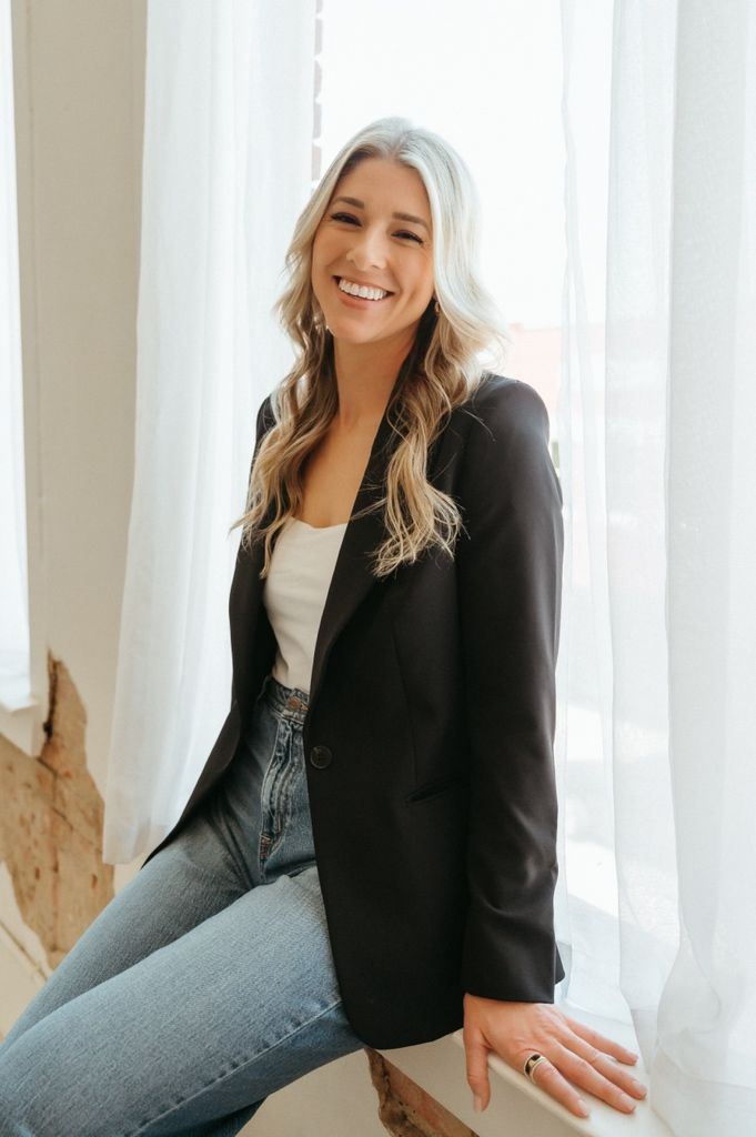 Smiling woman in black blazer and jeans poses by a bright window with sheer white curtains.
