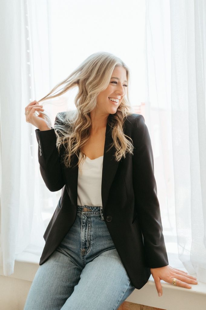 Cheerful blonde woman in black blazer and jeans playing with her hair near a sunlit window.