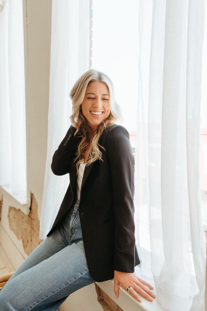 Smiling blonde woman in black blazer and jeans posing by sheer white curtains in bright natural light.