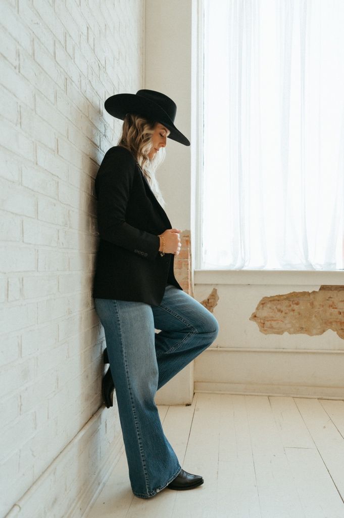 Woman in black blazer, wide-brim hat, and flare jeans leaning against white brick wall near window.