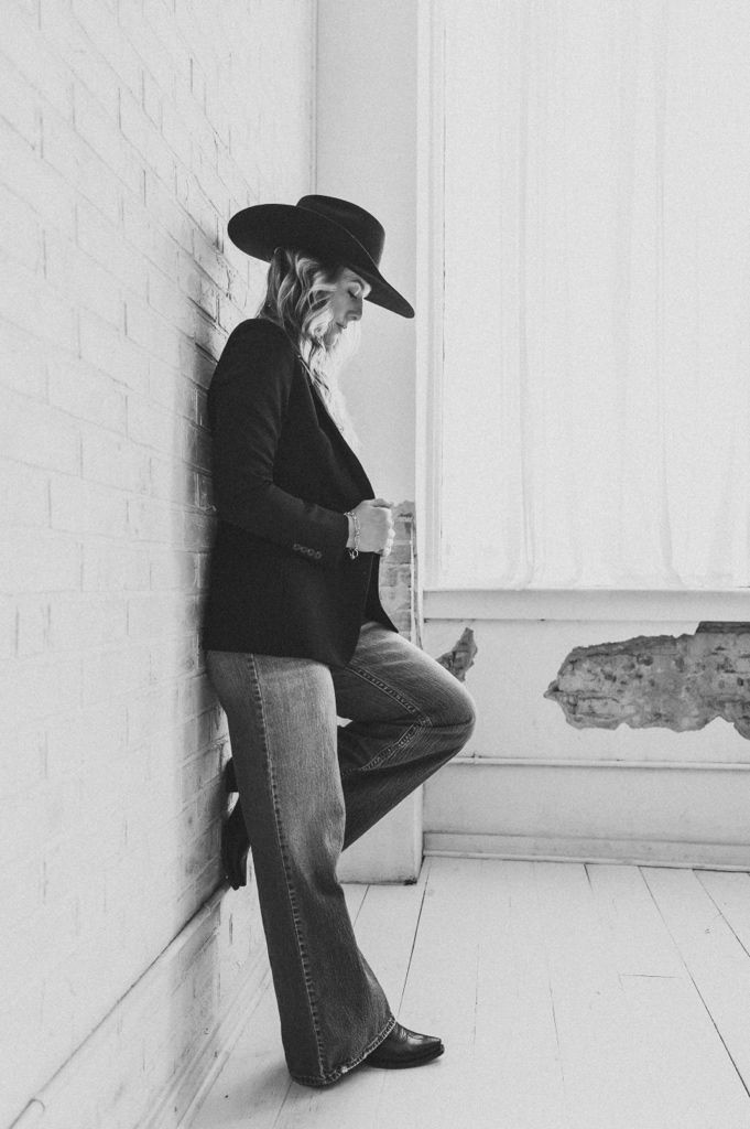 Black and white photo of a woman in a wide-brim hat, blazer, and flare jeans leaning against a white brick wall.