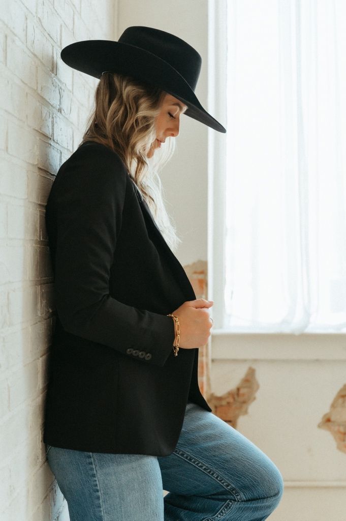 Woman in black wide-brim hat and blazer with jeans leans against white brick wall, looking down.
