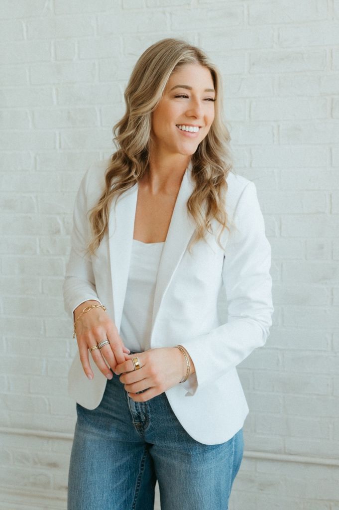 Smiling blonde woman in white blazer and jeans posing against a white brick wall.