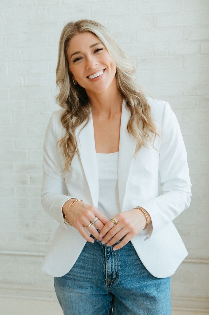 Smiling blonde woman in white blazer and jeans poses confidently against a white brick wall.