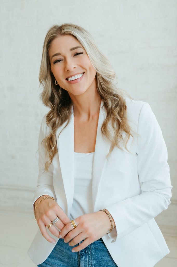 Smiling blonde woman in white blazer and jeans, wearing gold jewelry, posing confidently against a light background.