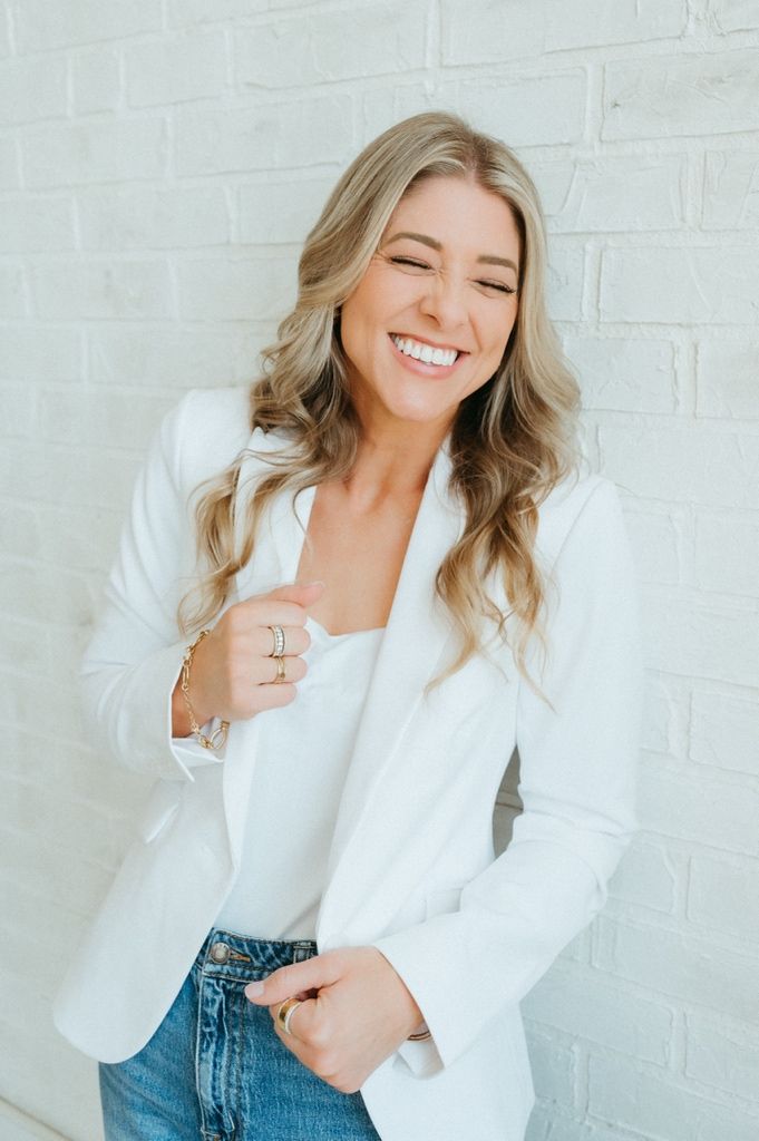 Smiling blonde woman in white blazer and jeans poses against a white brick wall wearing rings and bracelets.