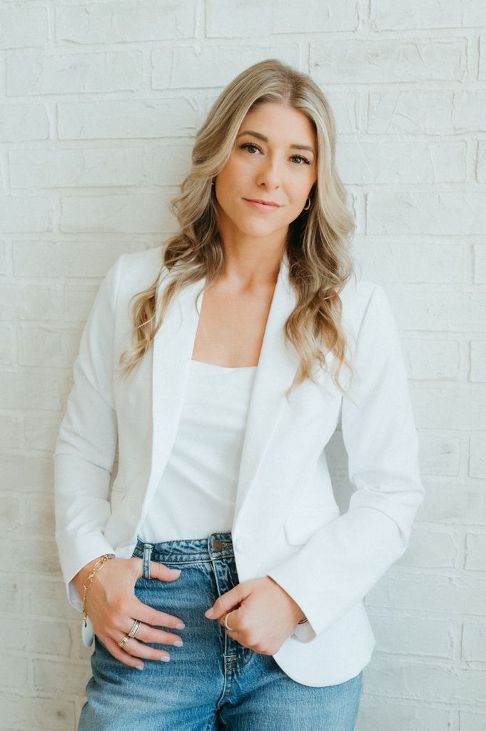 Woman in white blazer and jeans poses confidently against a white brick wall with long blonde wavy hair.
