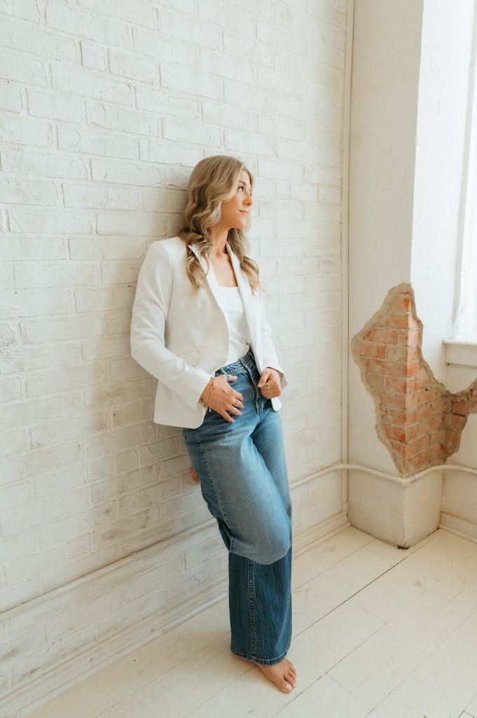 Woman in white blazer and wide-leg jeans leaning against a white brick wall in a bright, airy room.