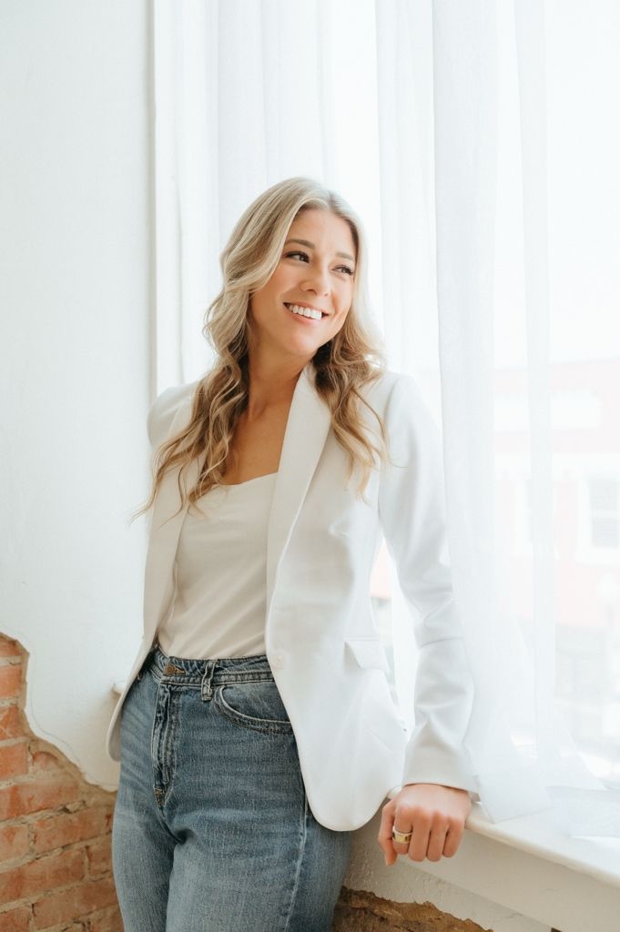 Smiling blonde woman in white blazer and jeans leaning against a window with soft natural light.