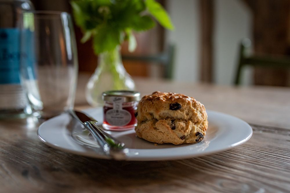 A golden brown scone sits on a white plate with a small jar of jam on a rustic wooden table.