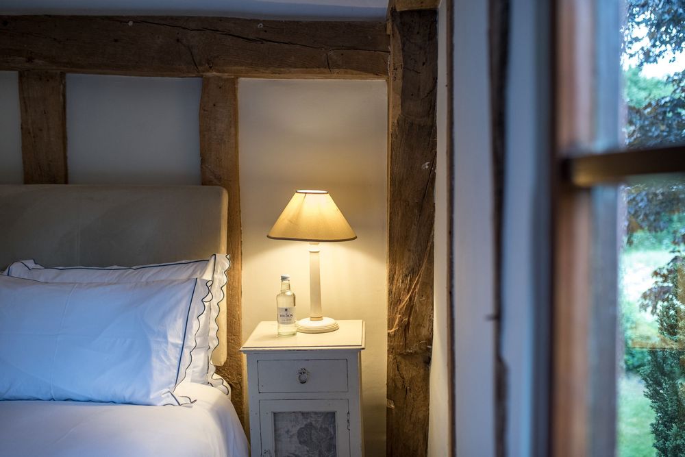 A cozy bedroom corner with exposed wooden beams, white bedding and a warm glowing table lamp on a nightstand.