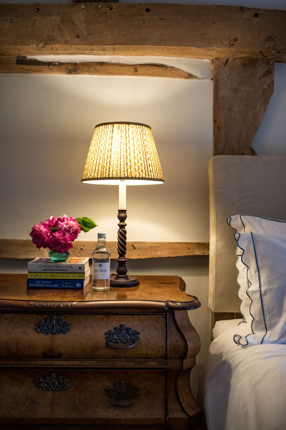 Cozy bedside table with traditional lamp, pink flowers, and books beneath rustic wooden beam ceiling.