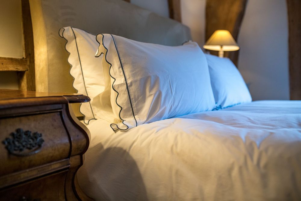Cozy bedroom detail showing white pillows with decorative trim and an antique wooden nightstand with warm lighting.