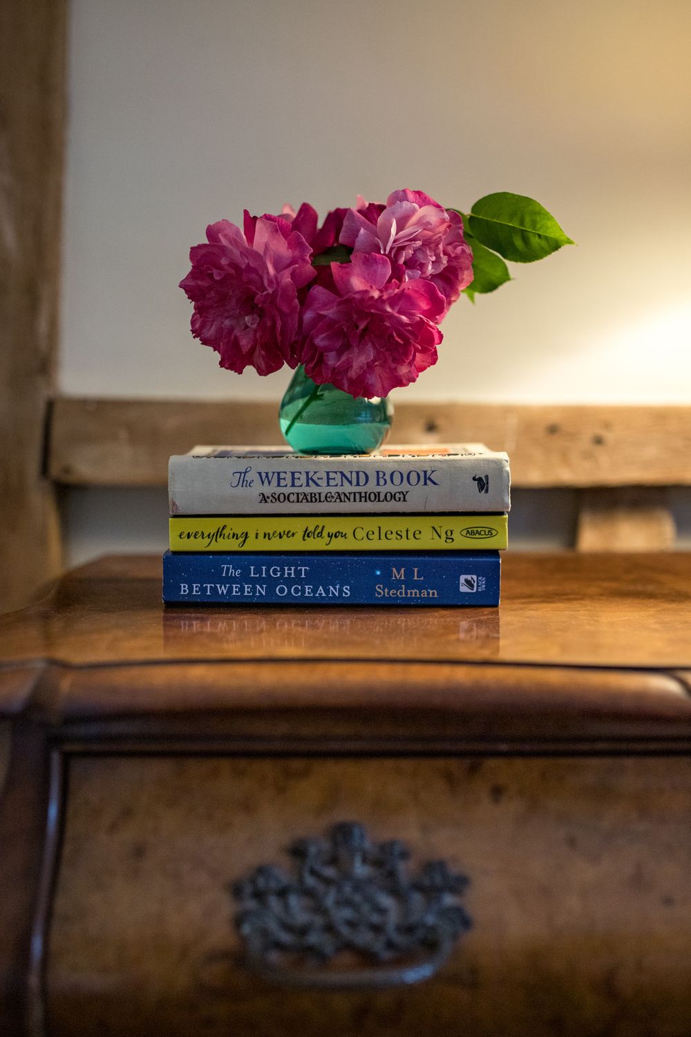 Pink roses in vase atop stack of novels on vintage wooden dresser with ornate handle.