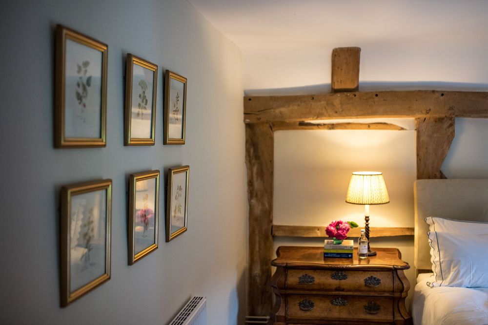 Rustic bedroom interior with framed botanical prints on wall and wooden beam accent above illuminated bedside table.