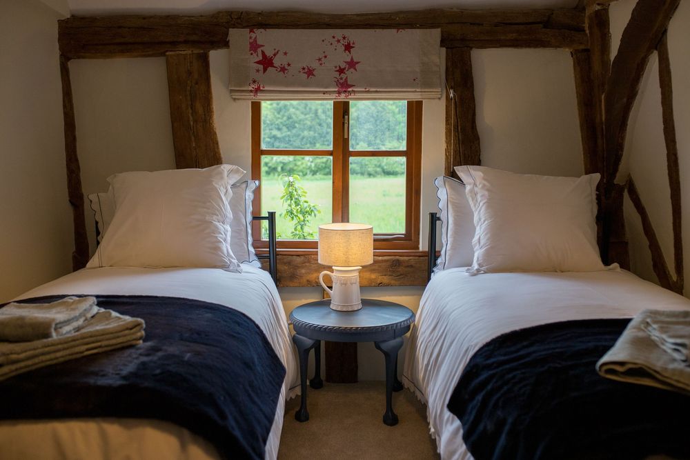 Cozy bedroom with twin beds dressed in white linens and navy blankets, featuring exposed wooden beams and a window.