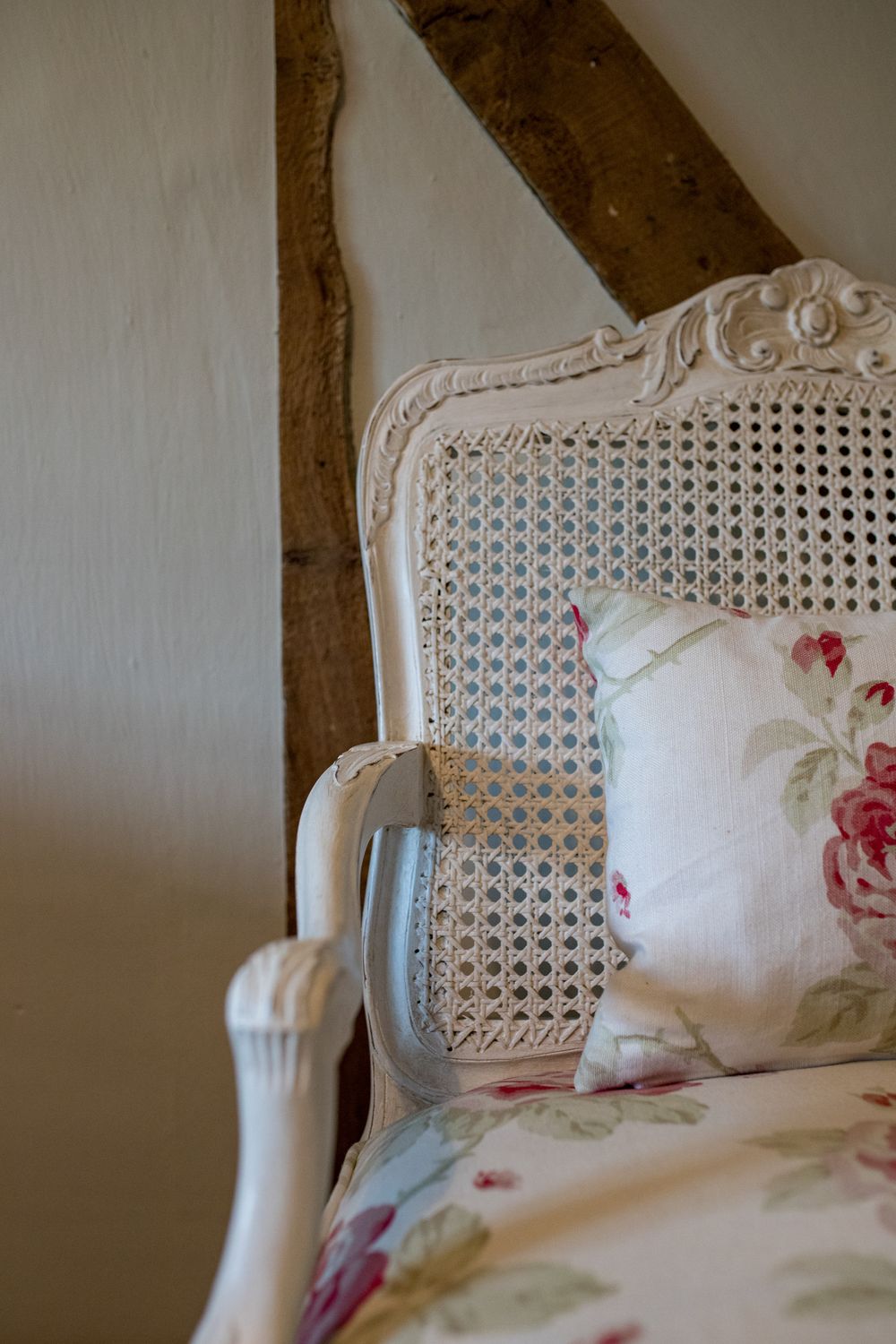 Close-up view of vintage cane chair with white painted frame and pink floral cushions against wooden beams.