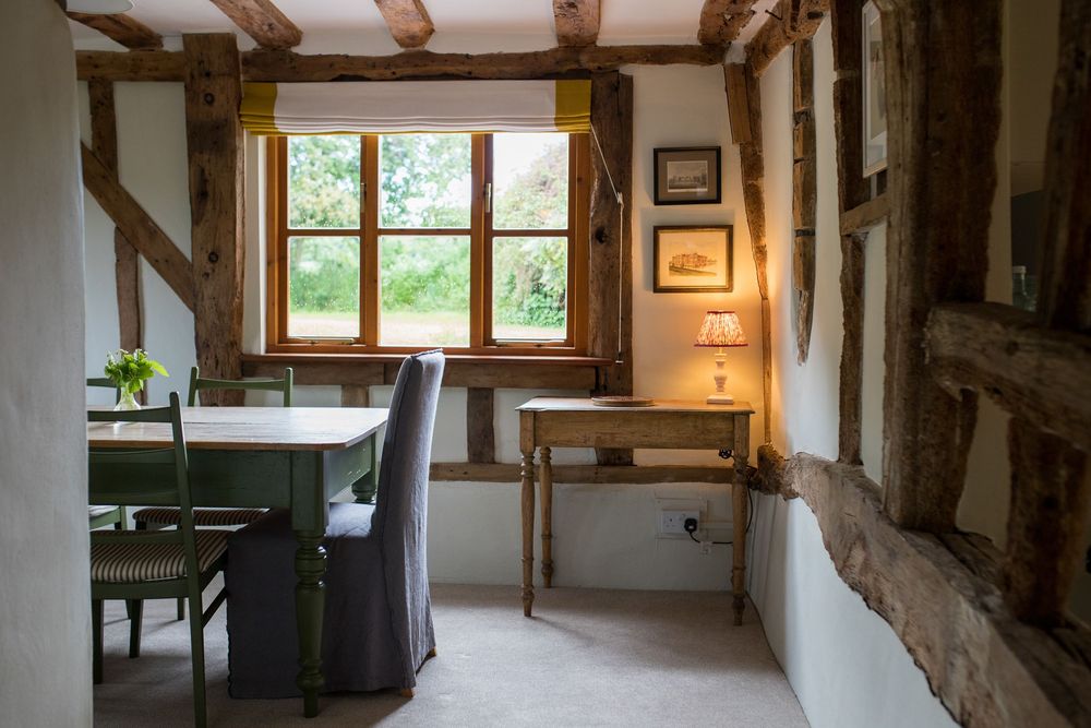 Rustic dining room with exposed wooden beams, white walls, and natural light streaming through windows in a cottage setting.