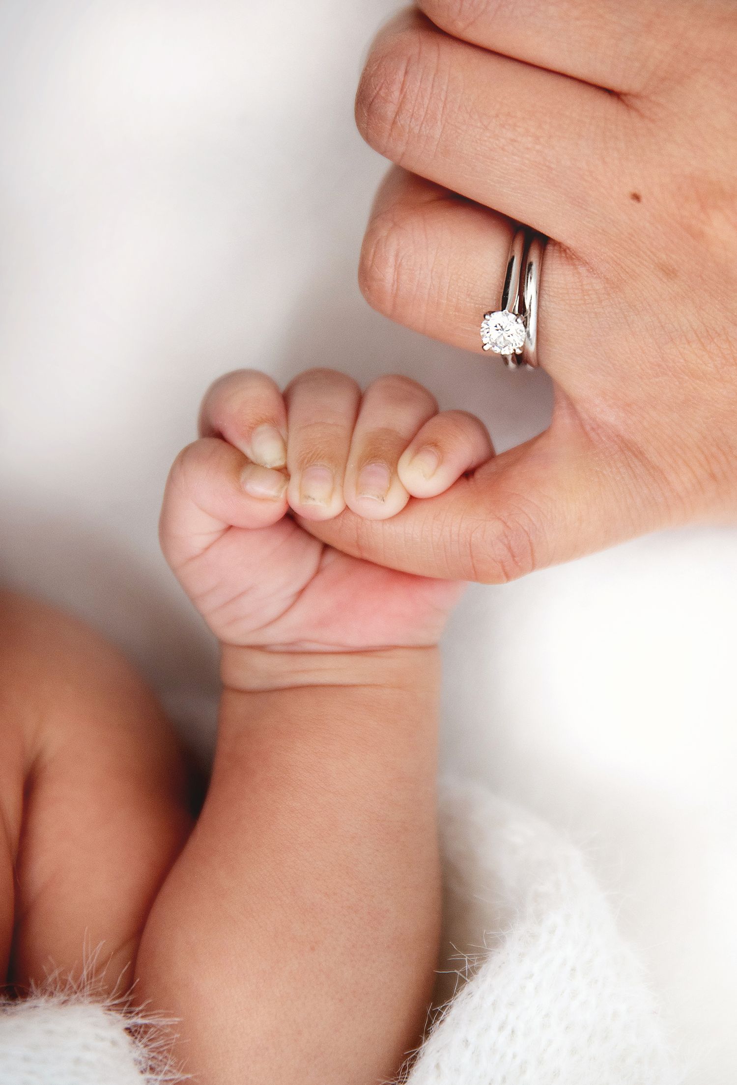 A newborn baby's tiny hand gripping an adult's finger wearing a diamond engagement ring. During a newborn photoshoot in Bexley