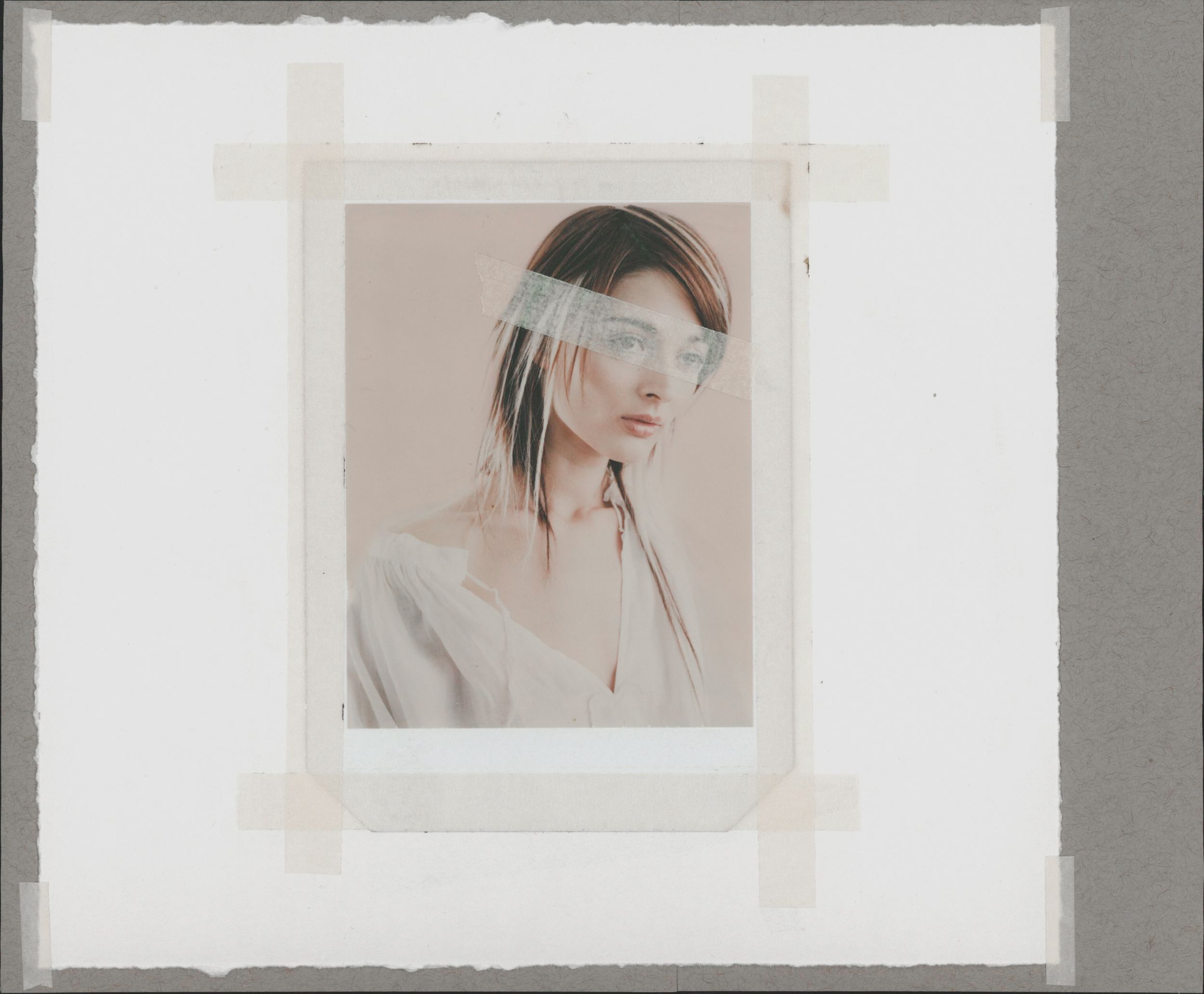 A woman with dark hair poses in a white top, photo taped to gray background with translucent strip across her eyes.