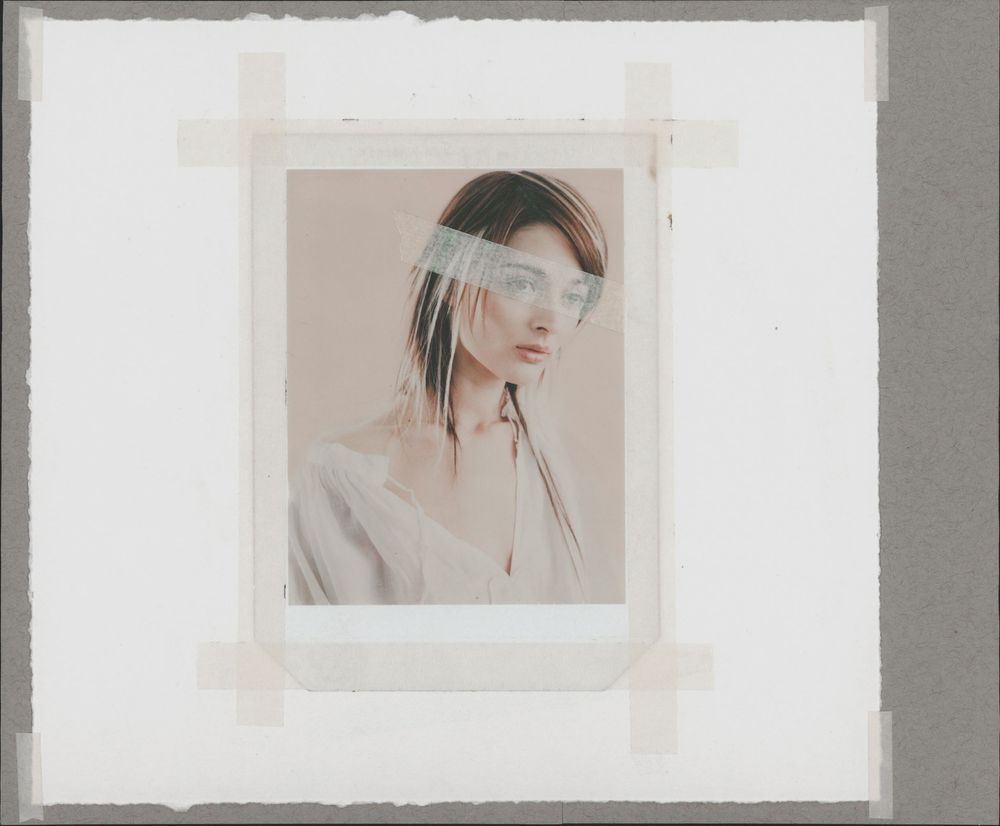 A woman with dark hair poses in a white top, photo taped to gray background with translucent strip across her eyes.