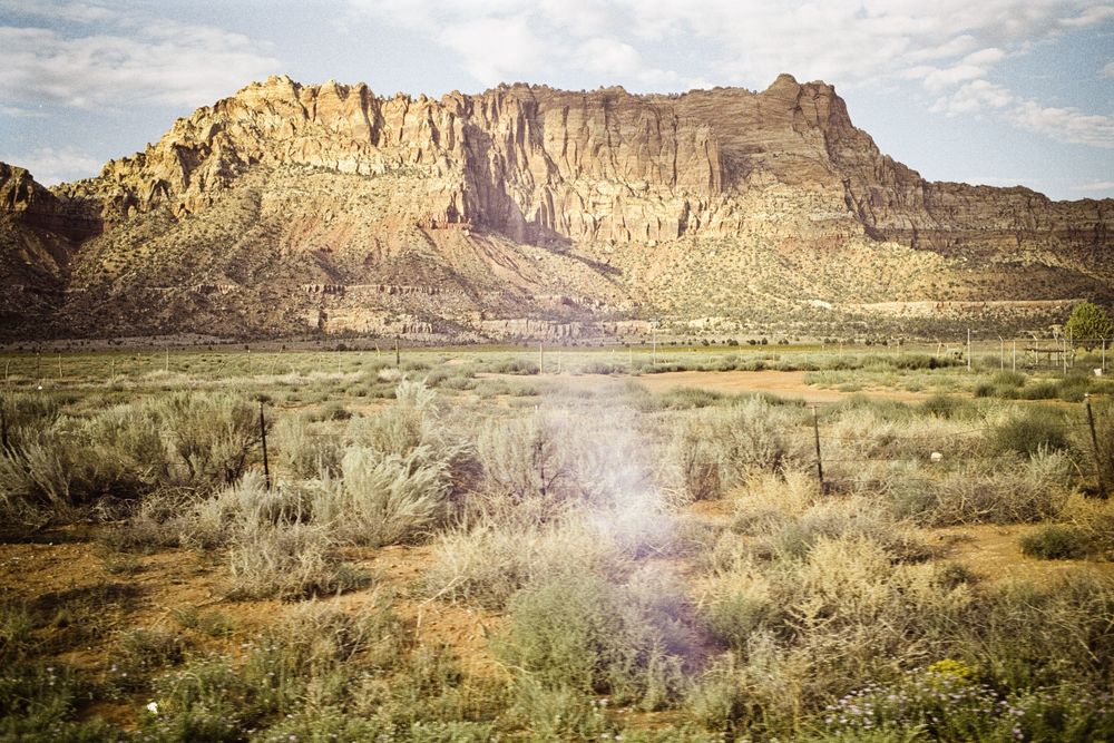Vast desert landscape with rugged sandstone cliffs, sagebrush plains, and a dusty haze rising under warm golden sunlight.
