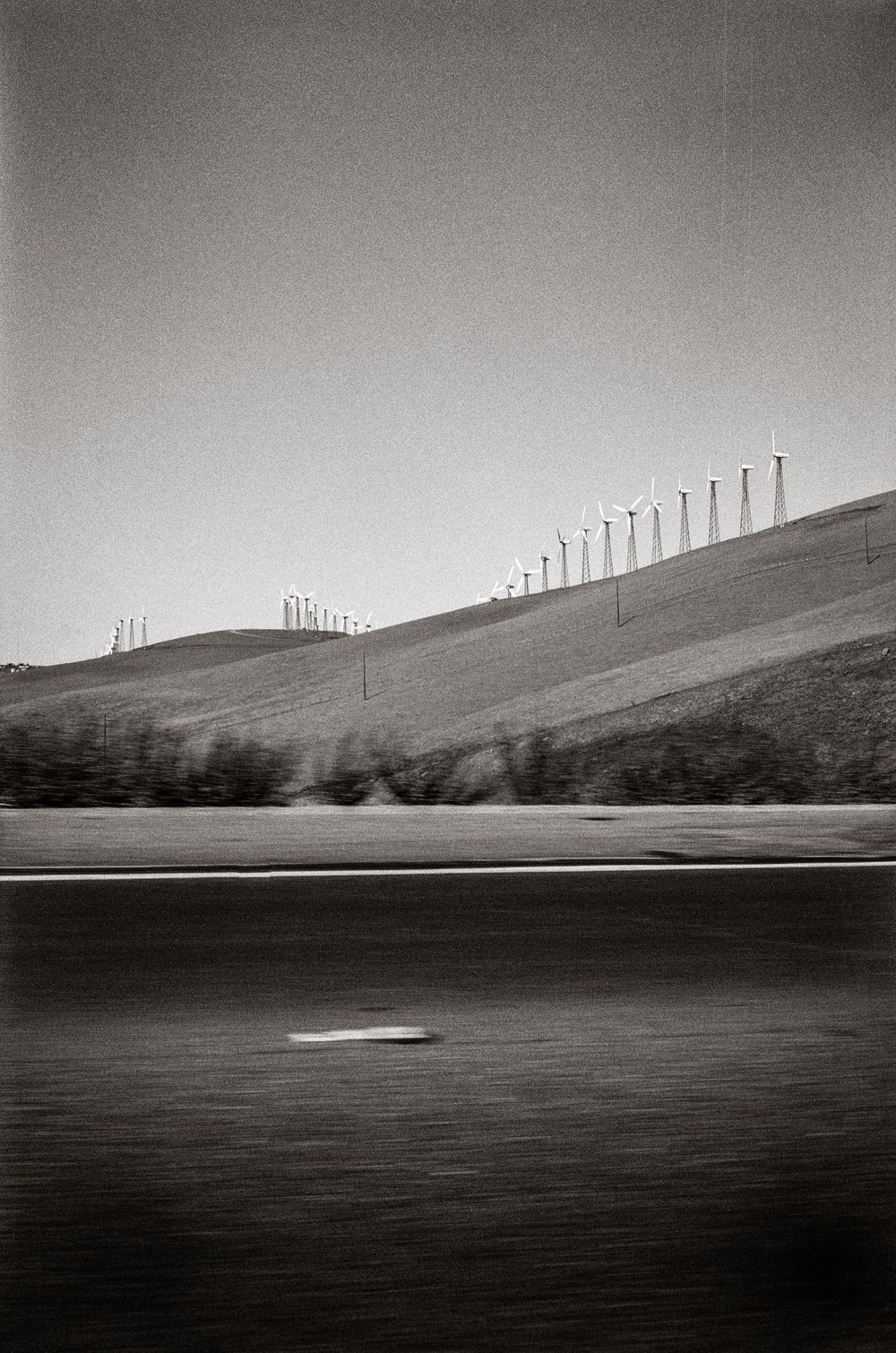 Black and white photo of wind turbines on rolling hills beside a blurred highway, captured from a moving vehicle.