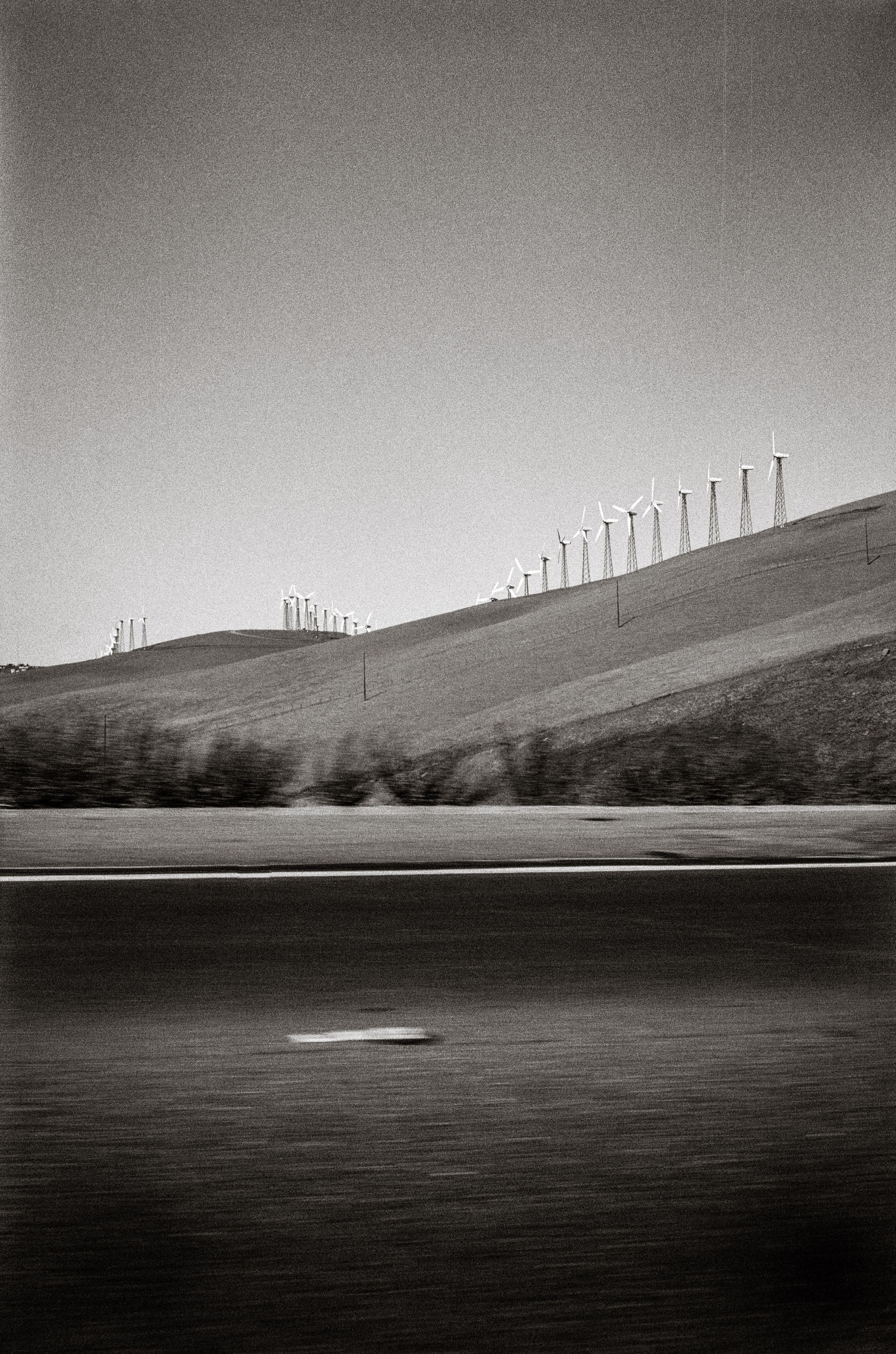 Black and white photo of wind turbines on rolling hills beside a blurred highway, captured from a moving vehicle.
